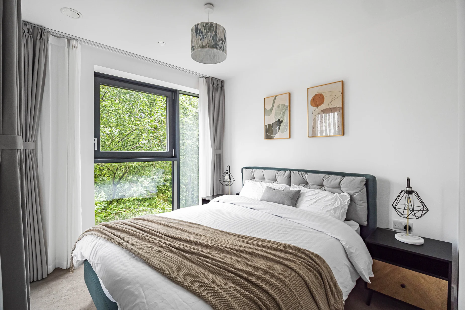 Neutral bedroom setting with layered cushions, taupe throw, bedside lamps and soft grey window dressing