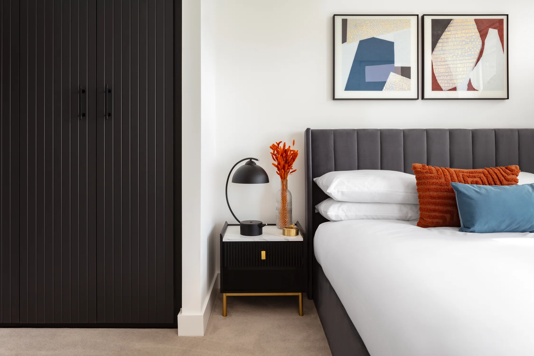 Bedroom styling detail with orange and blue cushions, framed artwork and black-and-brass bedside table